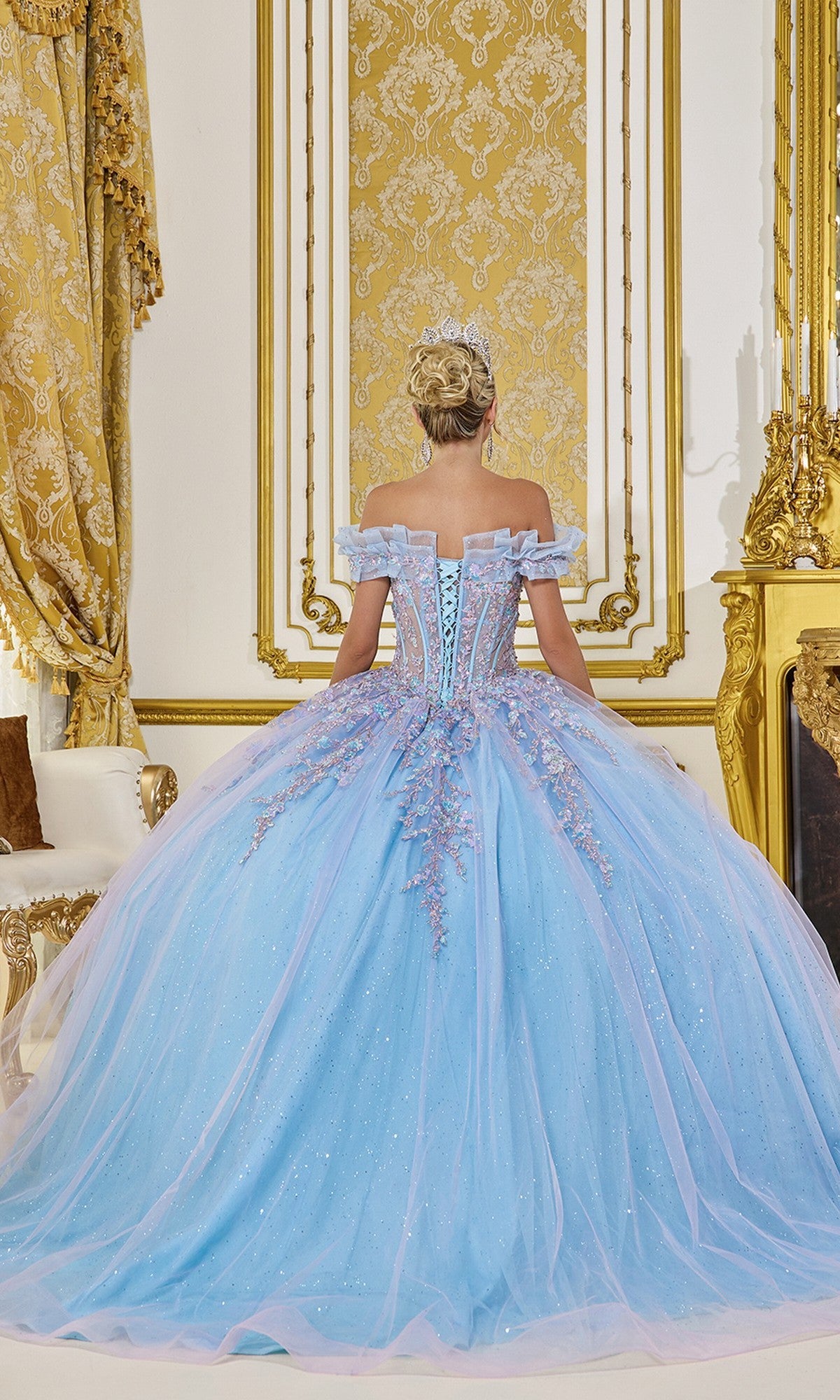 A woman in the Quinceanera Dress 5084 by Dancing Queen, an ornate blue off-the-shoulder gown, stands with her back to the camera in an opulent, gold-decorated room.